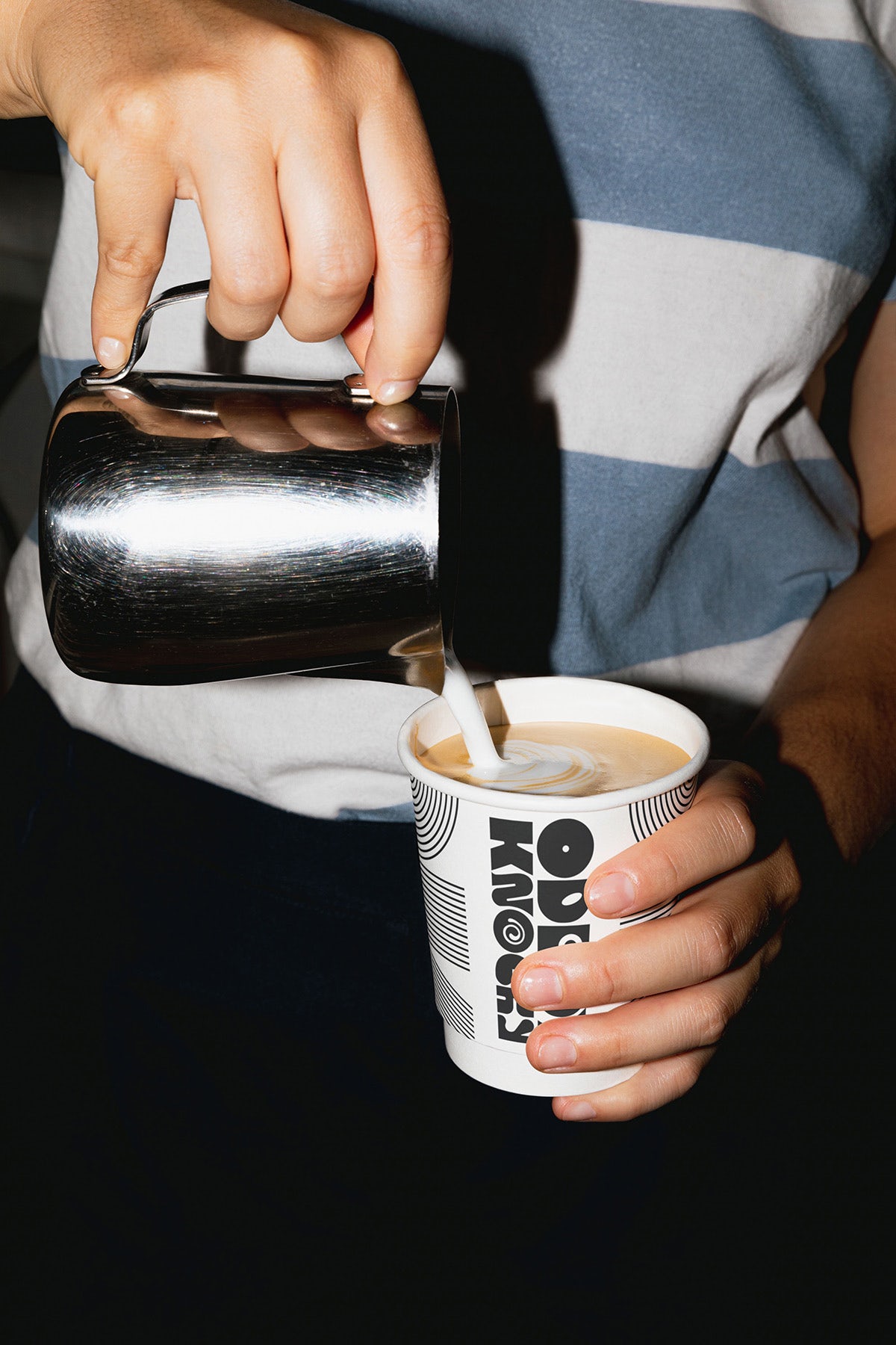 Photo showing a person pouring milk from a silver jug into a takeout cup of coffee decorated in Oddy Knocky's branding and multiline logo